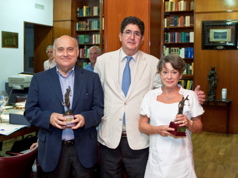 Los tres personas son felices, dos de ellos sostienen trofeos mientras la tercera observa. La escena se desarrolla en un ambiente de oficina o biblioteca, con estanterías llenas de libros y una mesa en el fondo.