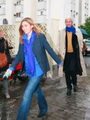 Dos personas caminan por una calle con casas blancas en el fondo. La persona de la izquierda lleva un abrigo azul y jeans, mientras que la de la derecha usa una chaqueta beige con un suéter amarillo. Ambas llevan bufandas azules.