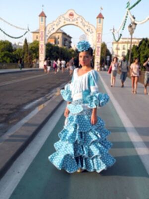Una mujer vestida con un traje de flamenca azul con lunares blancos y un tocado similar, camina por una calle con una arquería de entrada a un parque en el fondo.