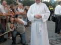 Un sacerdote vestido con un hábito blanco se encuentra en el centro de una calle, rodeado por personas que observan o toman fotos. La escena parece ser parte de una celebración o evento público, con edificios blancos y azules al fondo.