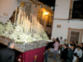 Una procesión religiosa con una imagen de la Virgen en un carro adornado con flores y velas. La gente observa atentamente, algunos llevando bolsas blancas. El ambiente es festivo y religioso en una calle con edificios de dos pisos.
