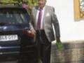 Un hombre en traje gris y corbata roja camina con un portafolios verde cerca de una BMW azul. La imagen muestra un ambiente formal, posiblemente en una ceremonia o evento importante.