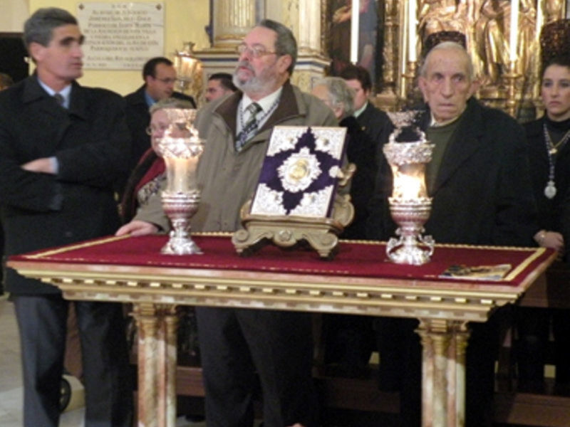 A una persona sentada en el centro, rodeada por otros individuos vestidos con trajes formales. En la mesa frente a ellos hay dos candelabros y un objeto decorativo con una insignia dorada. El fondo muestra detalles de una iglesia con estatuas y un altar.