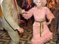 Una anciana vestida de rosa con un cinturón verde, baila con un hombre en traje. La escena parece ser una fiesta o evento formal, con otros invitados observando la danza.