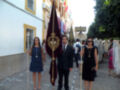Una imagen de una procesión religiosa con personas vestidas formalmente, entre ellas dos jóvenes que sostienen un escudo y una bandera. El entorno muestra edificios típicos de la ciudad, con una calle adoquinada y personas en el fondo.