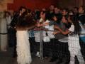 Una mujer con un vestido largo y capa de piel se encuentra entre una fila de personas detrás de barreras metálicas. La escena parece ser en un evento o fiesta, con gente esperando para saludar a la mujer.