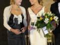 Dos mujeres sonríen mientras una sostiene un ramo de flores. Una lleva un vestido negro con detalles blancos y la otra, una falda blanca. Detrás de ellas, un hombre en traje negro y corbata esparcida. La escena parece ser una ceremonia o evento formal.