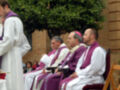Misa religiosa con sacerdotes vestidos de color blanco y morado sentados en sillas frente a un edificio con arcos.