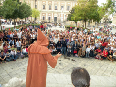 Una persona en un hábito marrón está dando una oración a una multitud de personas reunidas en un espacio público con edificios históricos al fondo.