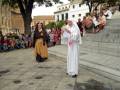Dos personas vestidas con trajes tradicionales de España, una en blanco y otra en marrón, caminan por un paso de piedra mientras una multitud observa desde las escaleras.