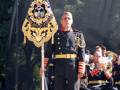 Un oficial militar sostiene una bandera ceremonial en un desfile. Detrás de él, otros oficiales y músicos se preparan para continuar la ceremonia.