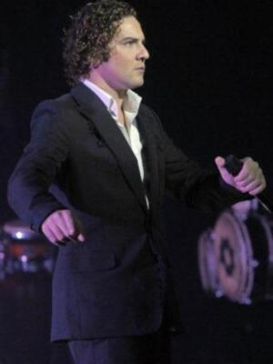 El hombre está en un escenario, con una camisa blanca y un abrigo negro. La iluminación es intensa, enfocándose en él.