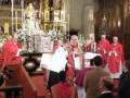 Imagen de una iglesia con un altar central, sacerdotes vestidos en rojo y blanco, fieles sentados en filas. El ambiente es reverente con detalles arquitectónicos y decoración religiosa.