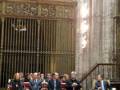 Un grupo de personas sentadas en bancos de madera en una sala amplia y alta, con detalles arquitectónicos ornamentados. La imagen muestra un ambiente formal y ceremonial, probablemente en una iglesia o edificio histórico.