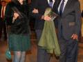 Una mujer con un vestido negro y tacones azules camina junto a dos hombres en traje, uno de ellos con un abrigo verde sobre el hombro.