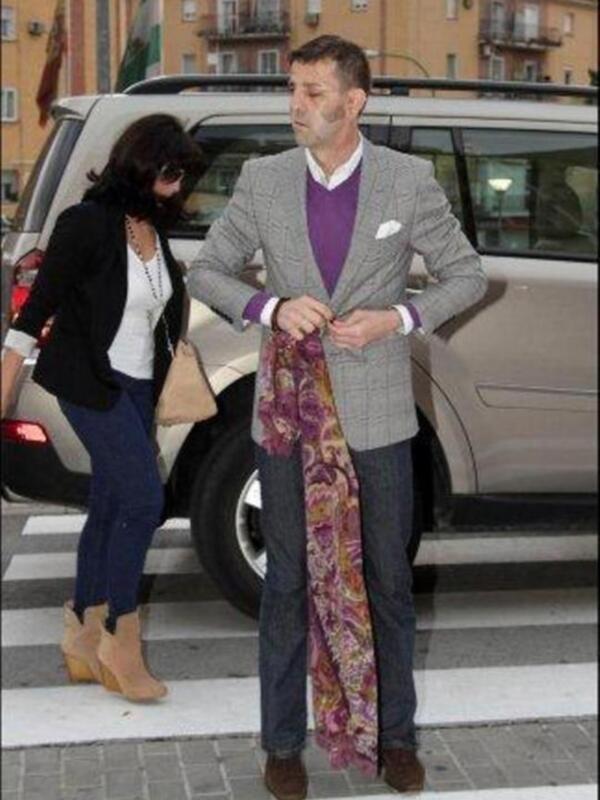 Un hombre y una mujer cruzan la calle en un entorno urbano. El hombre lleva un traje gris con una camisa morada y un pañuelo de colores. La mujer usa una chaqueta negra, una blusa blanca y pantalones azules. Detrás de ellos hay un vehículo beige y edificios en el fondo.
