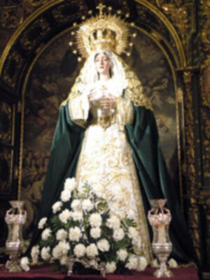 Virgen María en iglesia, vestida de blanco con capa verde y corona dorada. Fondo dorado con detalles artísticos. Base de flores blancas y velas en latón.