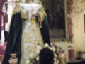 Imagen de una Virgen María vestida con trajes tradicionales y un velo blanco, rodeada de flores blancas y velas encendidas. En el fondo, un retrato de una familia con trajes históricos y un altar con velas.