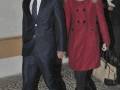 Dos personas caminan juntos, uno en traje formal y la otra con abrigo rojo y botas altas. La imagen parece ser de una celebridad o personaje importante, posiblemente en un evento formal.