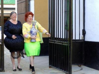 Dos personas vestidas con disfraces de personajes de dibujos animados caminan por una puerta metálica. La persona izquierda lleva un vestido negro y sandalias negras, mientras que la persona derecha usa una chaqueta amarilla con un vestido verde y sandalias negras. Ambas parecen estar disfrutando de la celebración.