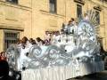 Un carro de carnaval plateado con detalles dorados, adornado con figuras y flores. La carroza está llena de personas vestidas de blanco, posiblemente bailando o participando en una celebración. La escena se desarrolla frente a un edificio de ladrillo, con ventanas y una barandilla visible.