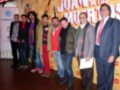 Un grupo de seis hombres posa en un escenario con una pancarta que dice "Juntos de los Muertos". Algunos llevan chaquetas y pantalones, mientras que otros usan camisas y jeans. La imagen está en una sala de conferencias con un fondo amarillo y azul.