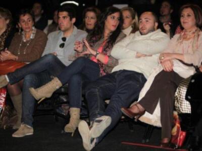 Un grupo de personas sentadas en una sala, algunas con pantalones cortos y otros con pantalones largos. Algunas personas llevan relojes, y algunas tienen sombrillas en sus manos. La imagen parece ser de una audiencia en un evento o conferencia.