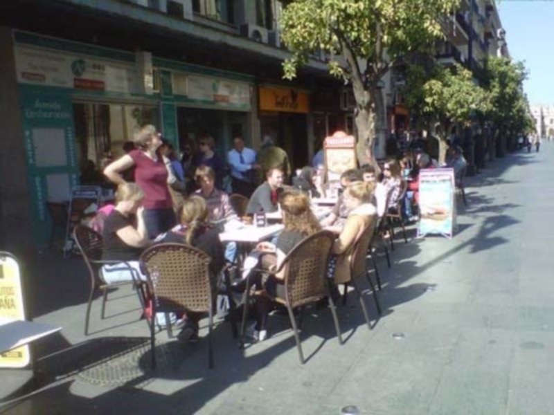Un grupo de personas disfruta de una comida al aire libre en un café. La calle está llena de gente sentada a mesas con sillas, y hay una fuerte presencia de sombrillas para protegerse del sol. La imagen muestra un ambiente animado y relajado, típico de una zona comercial o turística.