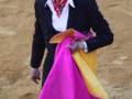 Un torero con un traje negro y una camisa blanca, sosteniendo una capa de color rosa y amarillo en la mano. La imagen se encuentra en un entorno de arena, probablemente durante una corrida de toros.