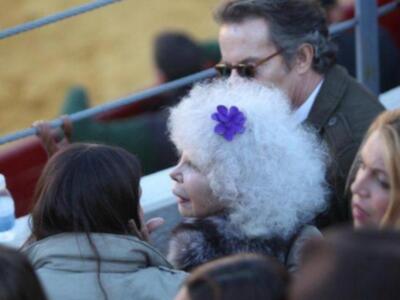 Una persona con peluca blanca y un flor en la frente, junto a otros individuos en una multitud.