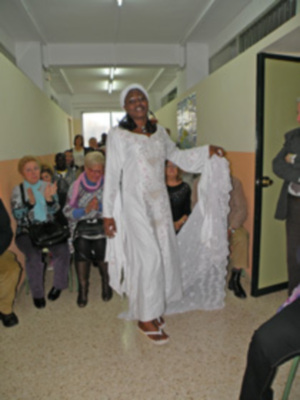 Una mujer con vestido blanco y sombrero negro sonríe mientras camina por un pasillo. Detrás de ella, se ven personas en diferentes poses y vestimentas. La imagen está iluminada con luces de techo y una puerta verde a la derecha.