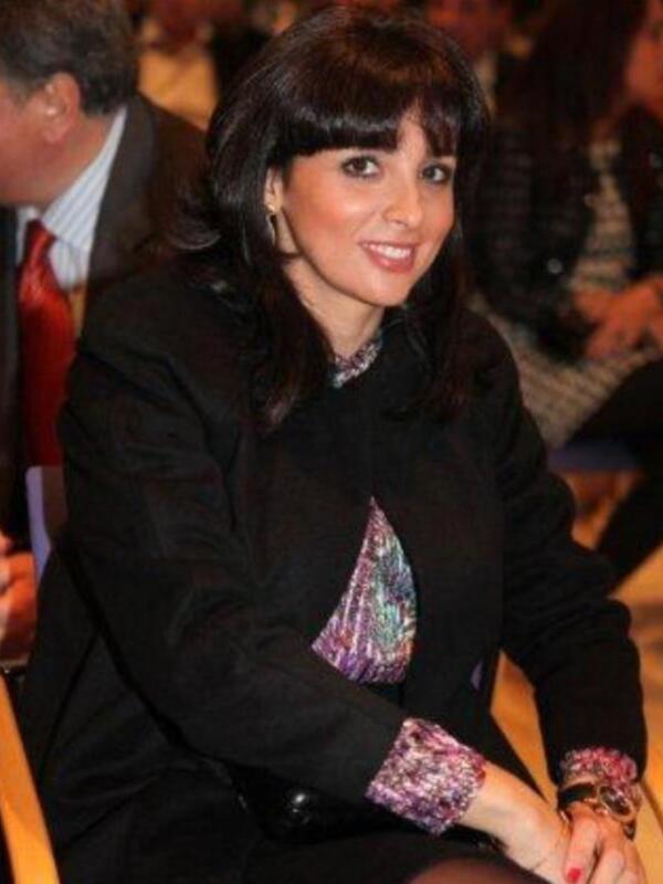 Una mujer con cabello oscuro corto y ondulado, lleva un traje negro con detalles brillantes en la manga. Está sentada en una sala de conferencias, con otros asistentes al fondo.