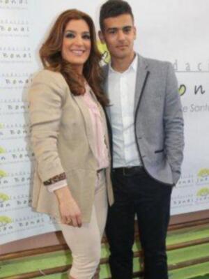 Una pareja posa frente a un fondo con el logo de una organización benéfica. La mujer lleva un traje beige con detalles en rosa y el hombre está con una camisa blanca y pantalones negros.