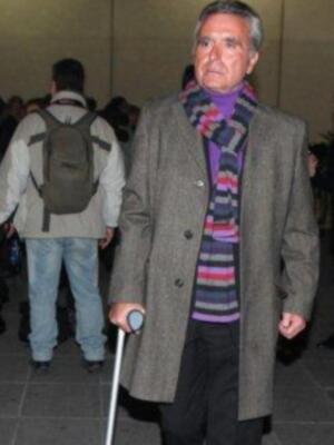 Un hombre de cabello gris, usando una chaqueta larga y un abrigo de color marrón claro. Lleva una camiseta con un cuello alto y un chal de colores vivos. También usa una muleta en la mano izquierda y un bolso grande en el hombro derecho. En el fondo, hay personas caminando y otros objetos como una mochila y un par de zapatos.
