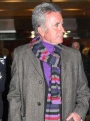 Un hombre con cabello gris corto y una chaqueta de tweed marrón. Lleva un abrigo largo con botones negros y una bufanda de colores brillantes. La imagen parece ser en un entorno urbano con luces de fondo.