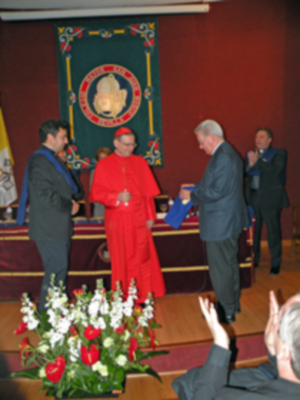 Un hombre en una capa roja sostiene un diploma mientras otro individuo lo felicita. La imagen muestra a varios personas en una ceremonia de reconocimiento, con un fondo que incluye un escudo y una bandera.