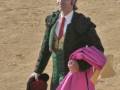Un torero en traje tradicional, con un chaleco verde y una falda rosa, sostiene su capote en la mano izquierda. El fondo es un paisaje desértico.