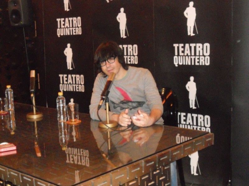 Una mujer con gafas está sentada en un escritorio, escribiendo algo en una libreta. A su lado hay botellas de agua y un bolígrafo. El fondo es negro con letras blancas que dicen "Teatro Quintero".