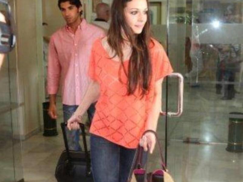 Mujer con chaqueta naranja y pantalones azules, caminando con una maleta. Hombre detrás de ella, también con chaqueta naranja y pantalones azules. Ambos caminan por un pasillo con puertas de cristal y espejos en el fondo.