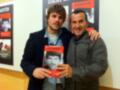 Dos hombres sonrientes posan con un libro titulado "Dani Martín" en una sala de exposiciones.