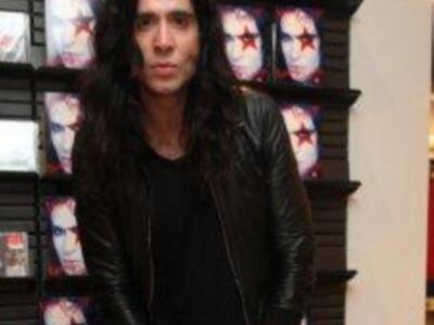 Un hombre con cabello largo y rojo, vestido con una chaqueta de cuero negra, posa frente a estanterías llenas de revistas con portadas rojas y blancas.