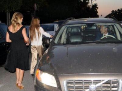 Dos mujeres caminan hacia un coche negro mientras un hombre se sienta en el asiento delantero. La escena parece ser de una celebridad o evento formal.