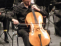 Un músico toca el violonchelo en un escenario de orquesta, con otros músicos al fondo.