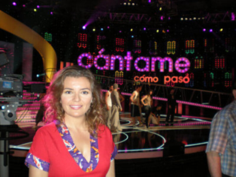 Una mujer con vestido rojo y flores posa en un escenario con luces y pantallas de fondo.