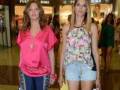 Dos mujeres caminan por un centro comercial, una con camiseta rosa y pantalones de chándal azules, la otra con una camiseta estampada y shorts cortos. En el fondo se ven otros clientes y un escaparate de tienda.