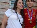 Una pareja en un evento deportivo, con la mujer vestida de blanco y el hombre con una camiseta de fútbol de España con una medalla dorada.