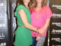 Dos mujeres posan juntas en un evento, uno con vestido verde y la otra con camisa rosa. Fondo con banners de Aromas.