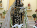 A una Virgen vestida con un manto blanco y un vestido negro adornado con detalles dorados. La Virgen está rodeada de flores blancas y rosadas, y hay un altar con velas en el fondo. La escena parece ser parte de una celebración religiosa o una procesión.