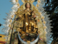 Imagen de una Virgen María con un vestido dorado y joyas, rodeada por ángeles y símbolos religiosos. Se encuentra en un entorno con edificios de colores y una clara luz del día.