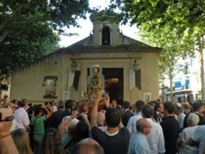 Una multitud congregada en torno a una imagen venerada, con un pequeño edificio de madera y una campana en el techo. La imagen central muestra a una Virgen con un niño, rodeada de fieles que la saludan y alzan sus manos en oración. La escena se desarrolla bajo un cielo despejado, con árboles y edificios alrededor.